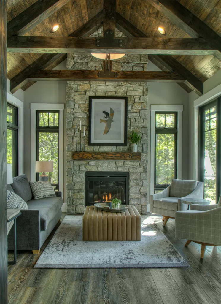Cozy lakeside living room with stone fireplace, timber‑beam ceiling, and large windows framing wooded views; gray sofa, plaid armchairs, and tufted ottoman create a welcoming cabin atmosphere.