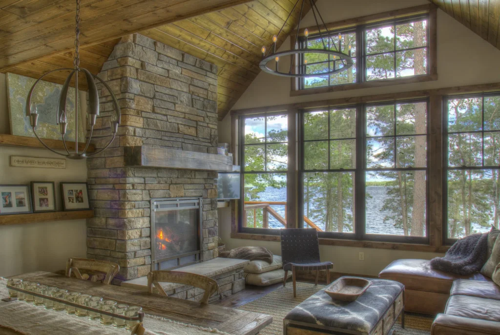 Inviting lakeside living room with stone fireplace, timber‑beam ceiling, and expansive windows overlooking the water; cozy seating and rustic wood accents create a relaxed cabin atmosphere.