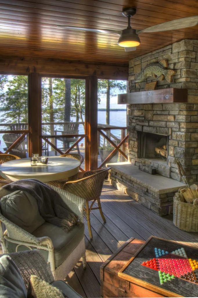 Screened lakeside porch with stone fireplace, wicker seating, and wood ceiling; cozy table and chairs overlook the water through large windows, creating a warm, inviting cabin retreat.