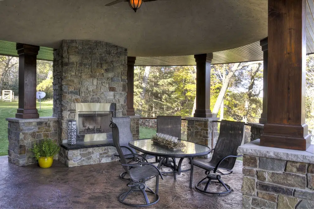 Covered lakeside patio with stone fireplace, round glass dining table, and cushioned chairs; wood columns frame views of trees and lawn, creating a cozy outdoor retreat for Minnesota lake homes.