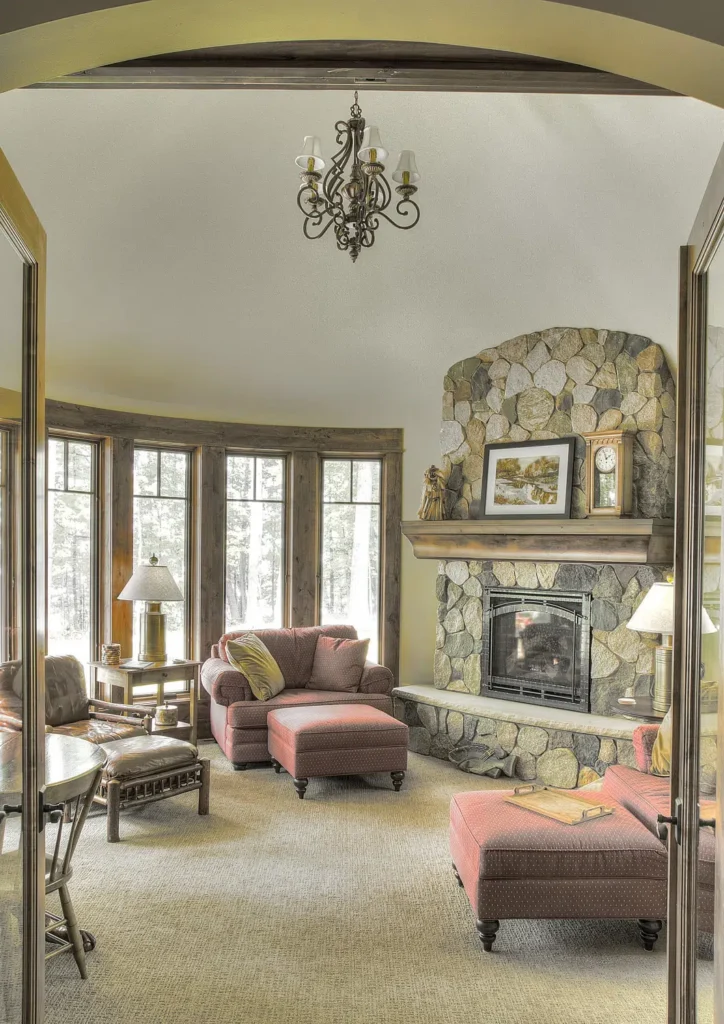 Inviting lakeside living room with stone fireplace, arched ceiling, and large windows framing forest views; cozy armchairs and ottomans create a comfortable retreat with rustic elegance.